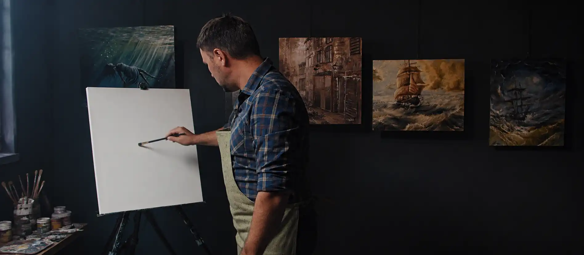 An artist standing before a blank canvas, finished paintings hanging on the wall behind him