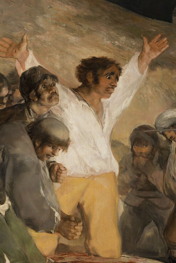 The man in a white shirt and yellow trousers kneeling with his arms flung wide, facing the rifles